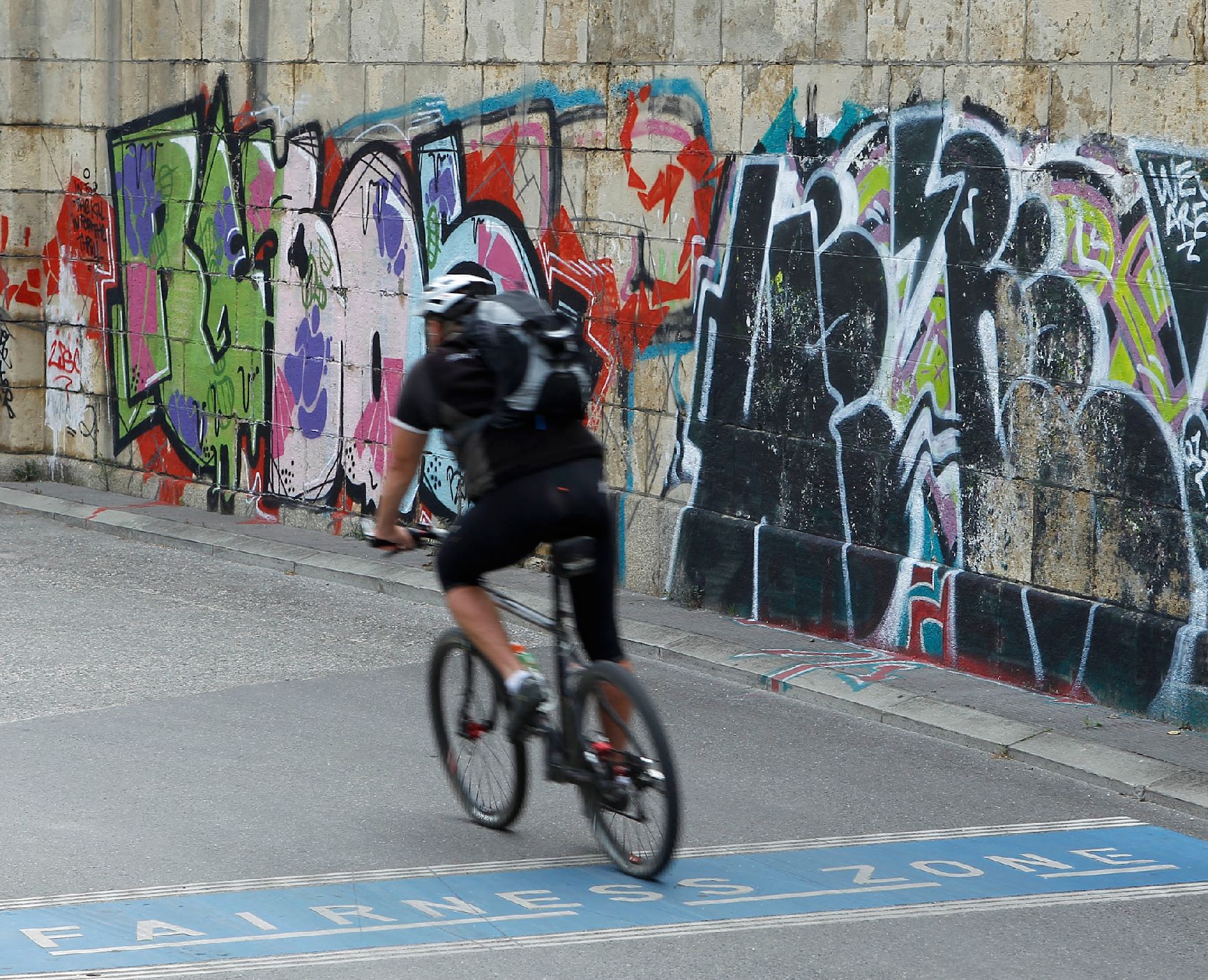 Ein Fahrradfahrer auf einem Fairness Zone - Radweg am Donaukanal. Im Hintergrund Graffitis an einer Mauer. Schlagworte: Fahrrad, Fahrzeug, Graffiti, Helm, Kunst, Mann, Mensch, Rucksack, Stadtleben, Verkehr