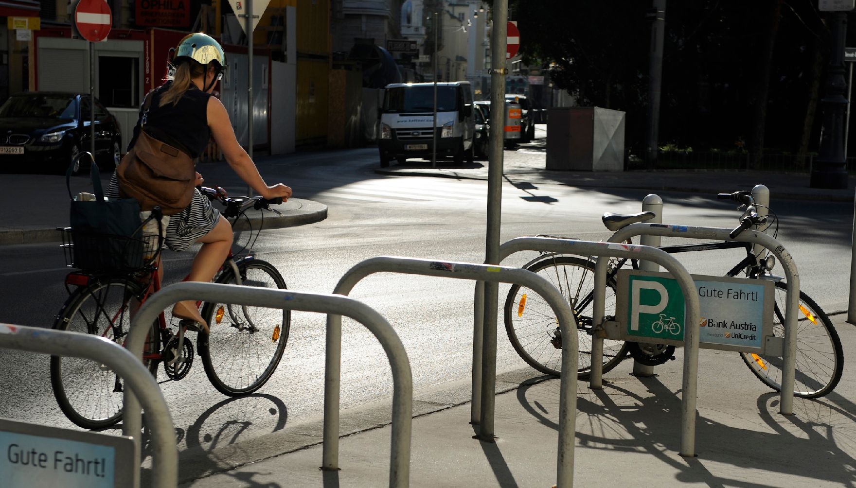Eine Fahrradfahrerin radelt an einem Radparkplatz vorbei. Schlagworte: Fahrrad, Fahrzeug, Helm, Frau, Mensch, Stadtleben, Straße, Verkehr