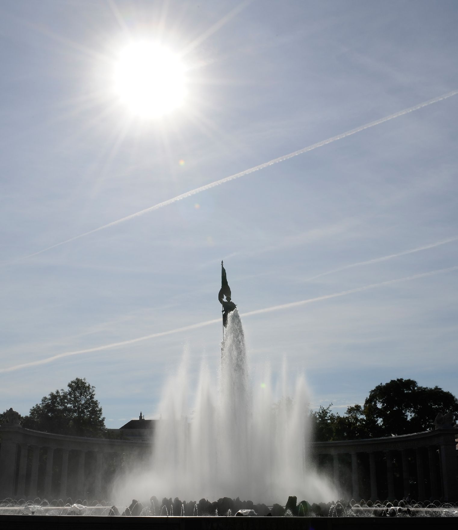 Der Hochstrahlbrunnen am Schwarzenbergplatz im Gegenlicht. Schlagworte: Brunnen, Himmel, Sonne, Stadtlandschaft, Statue, Wasser, Wolken