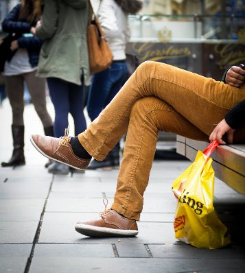 Eine Person mit Einkaufstasche. Schlagworte: Beine, Einkaufstasche, Füße, Hose, Menschen, Schuhe, Sitzbank, sitzen, Wirtschaft
