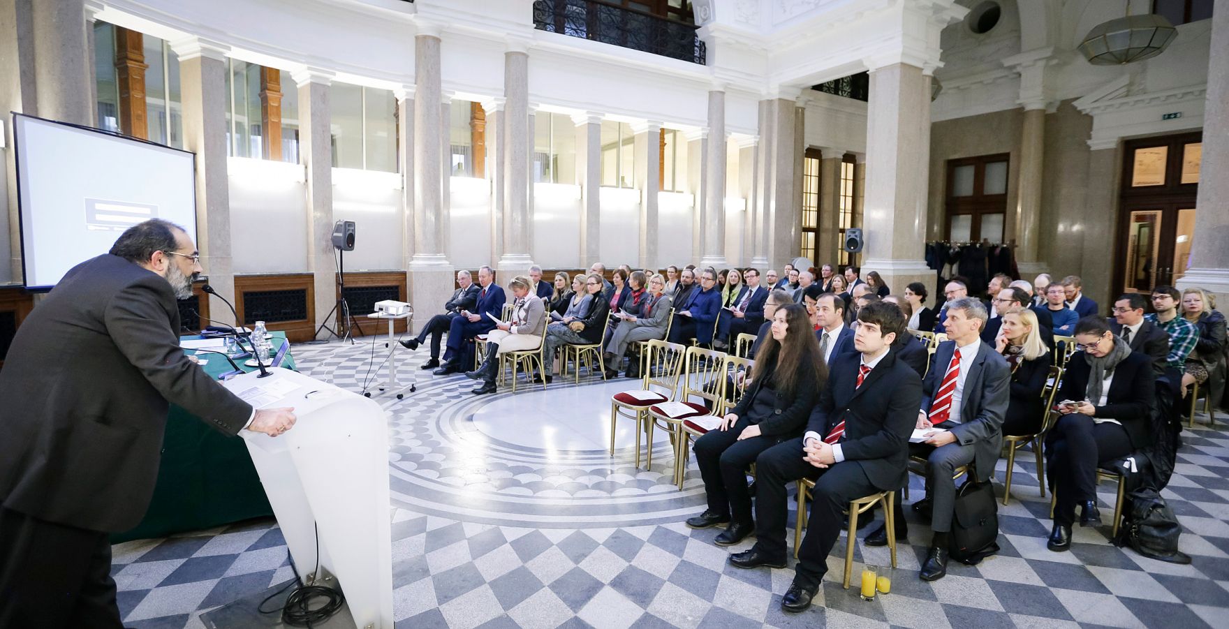 Am 7. M&auml;rz 2016 fand in Kooperation mit dem Dachverband der Verwaltungsrichter eine Vortrags- und Diskussionsveranstaltung mit Dr. Viktor Kreuschitz, Richter am Gericht des Gerichtshofes der Europ&auml;ischen Union, im Kassensaal des Amtsgeb&auml;udes Hohenstaufengasse 3, statt. Links im Bild Priv.-Doz. Alexander Balthasar bei der Begr&uuml;&szlig;ung, 2. Reihe rechts: Sen.Pr&auml;s. des VwGH Dr. Martin K&ouml;hler.