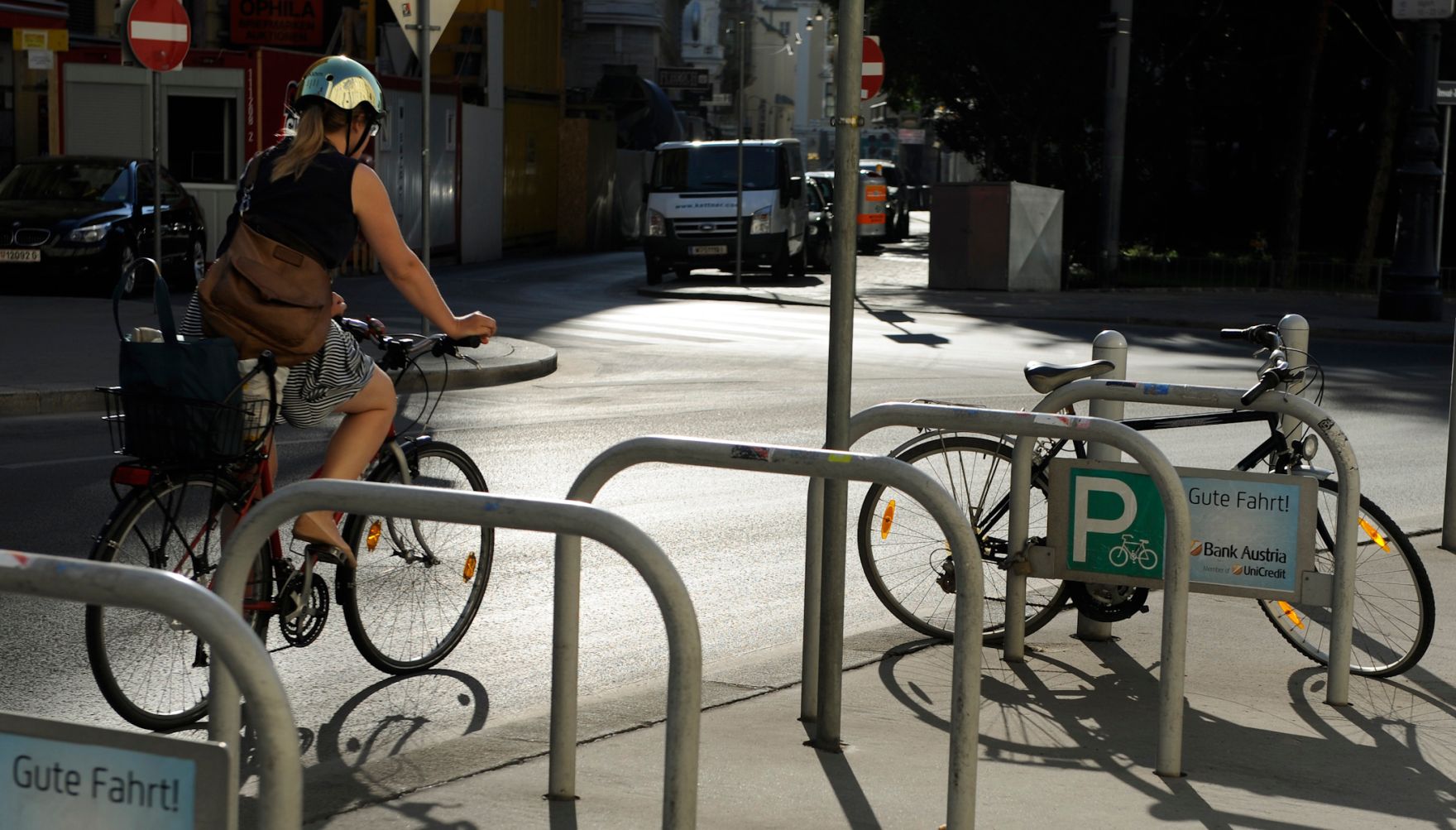 Eine Fahrradfahrerin radelt an einem Radparkplatz vorbei. Schlagworte: Fahrrad, Fahrzeug, Helm, Frau, Mensch, Stadtleben, Stra&szlig;e, Verkehr