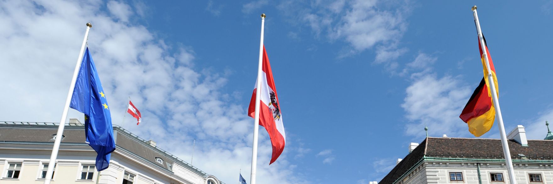 Die europ&auml;ische Fahne weht neben den Fahnen &Ouml;sterreichs und Deutschlands im Wind. Schlagworte: Bundeskanzleramt, Fahnen, Flaggen, Himmel, Masten, Staatsbesuch, Wolken