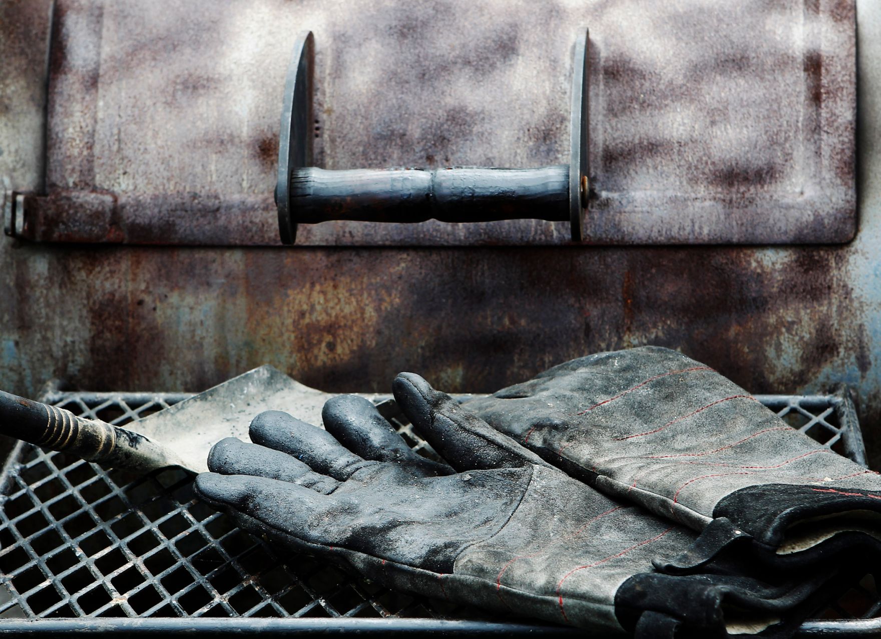 Ein Paar schmutzige Arbeitshandschuhe und eine kleine Schaufel liegen vor einem rostigen Griller. Schlagworte: Gitter, Metall, Rost