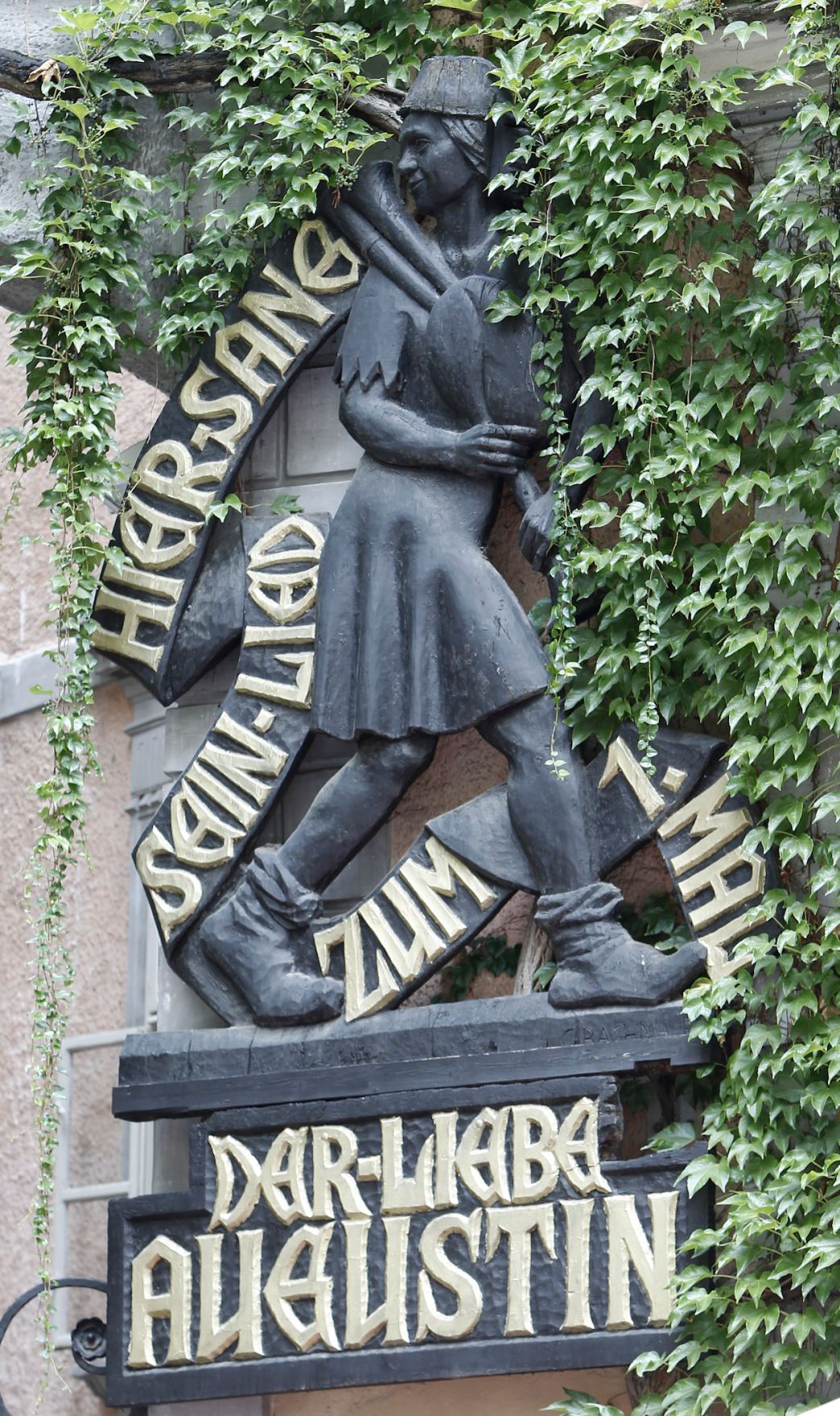 Holzfigur des "lieben Augustins" umrangt von Efeu. Schlagworte: Beschriftung, Denkmal, Stadtlandschaft, Statue