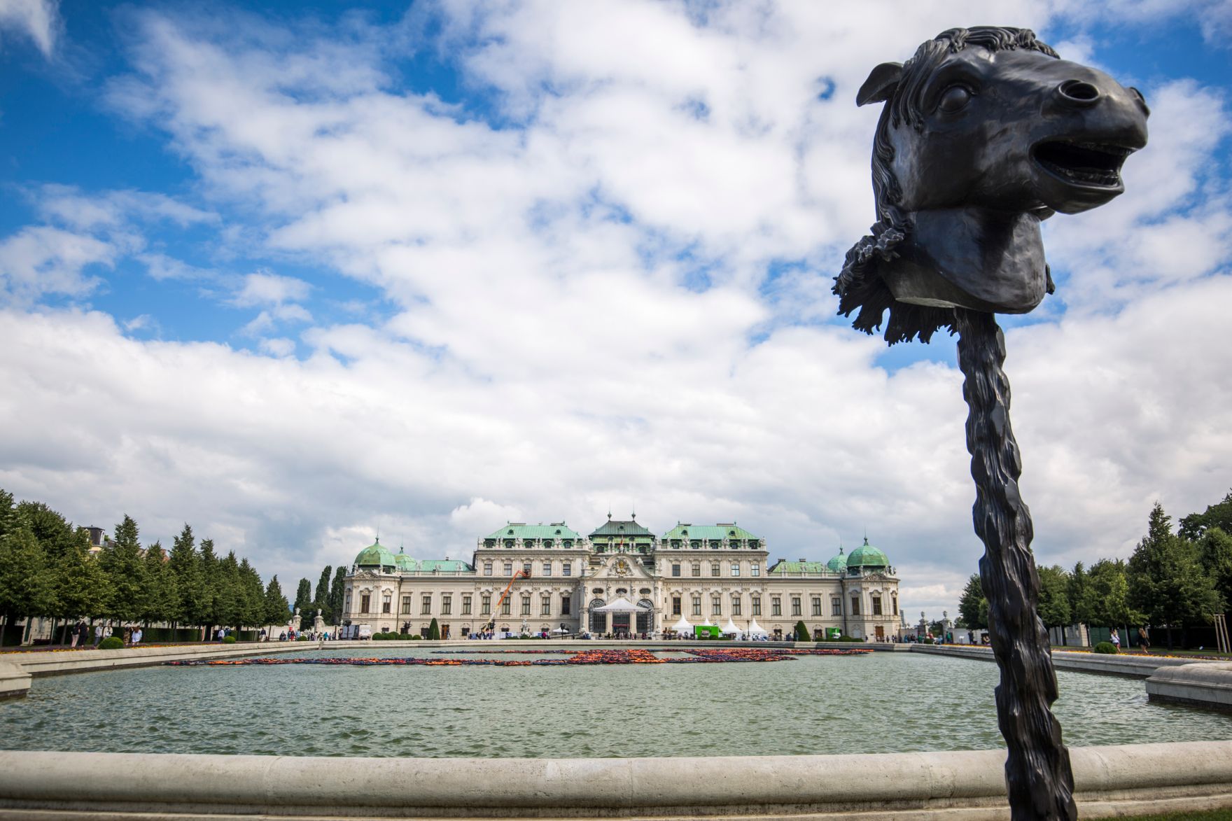 Pferdestatue des chinesischen K&uuml;nstlers Ai Weiwei, mit dem Schloss Belvedere im Hintergrund und Installation der Schwimmwesten von Fl&uuml;chtlingen im Wasser. Schlagworte: Ai Weiwei, Architektur, Ausstellung, Belvedere, F Lotus, Geb&auml;ude, Himmel, Kunst, K&uuml;nstler, Pferd, Sehensw&uuml;rdigkeit, Schloss, Schwimmwesten, Statue, Teich Tierkreiszeichen, Wasser, Wolken