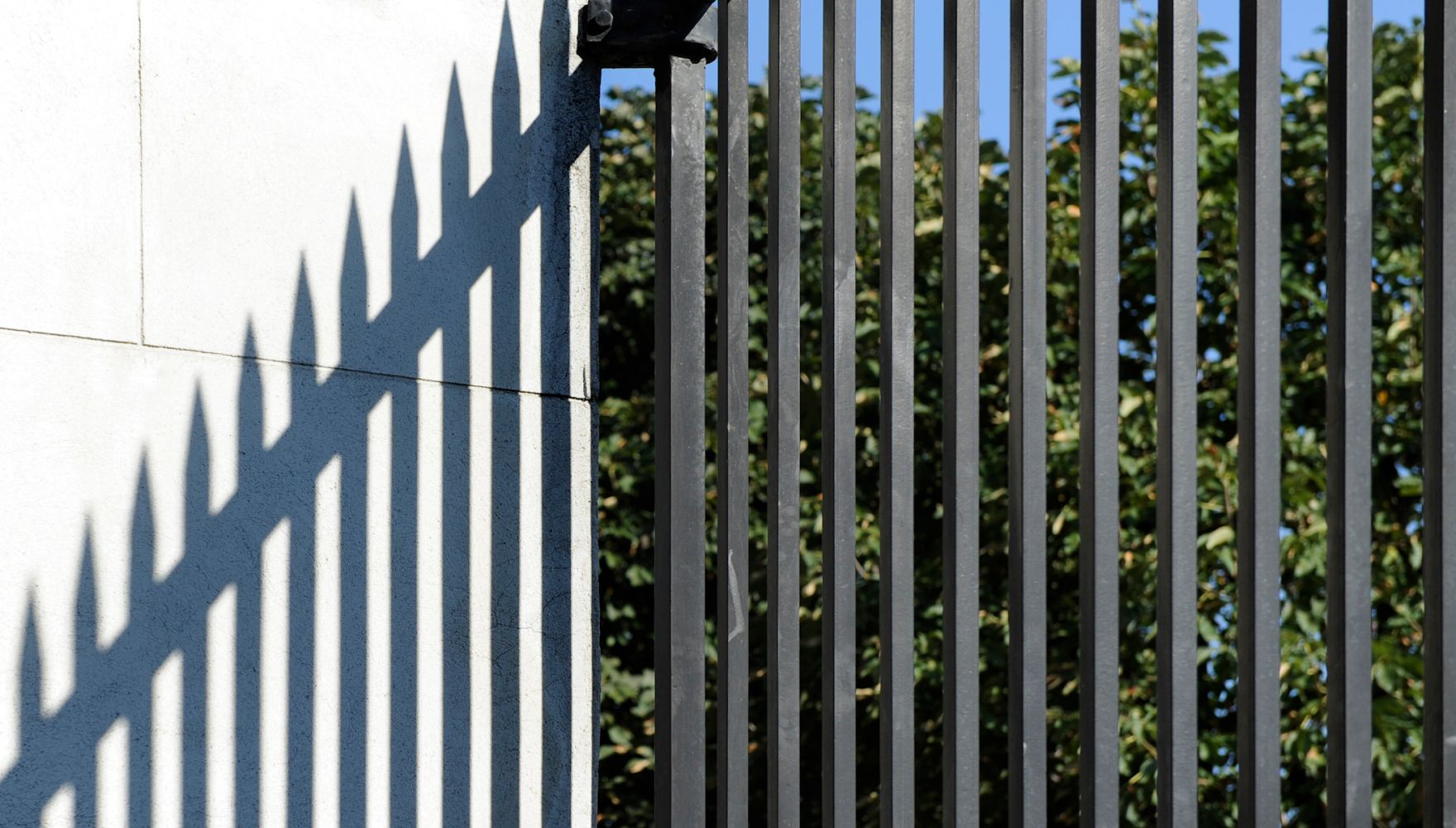 Ein Zaun wirft seinen Schatten an eine Wand. Schlagworte: Gitter, Mauer, Schatten, Stadtlandschaft, Zaun