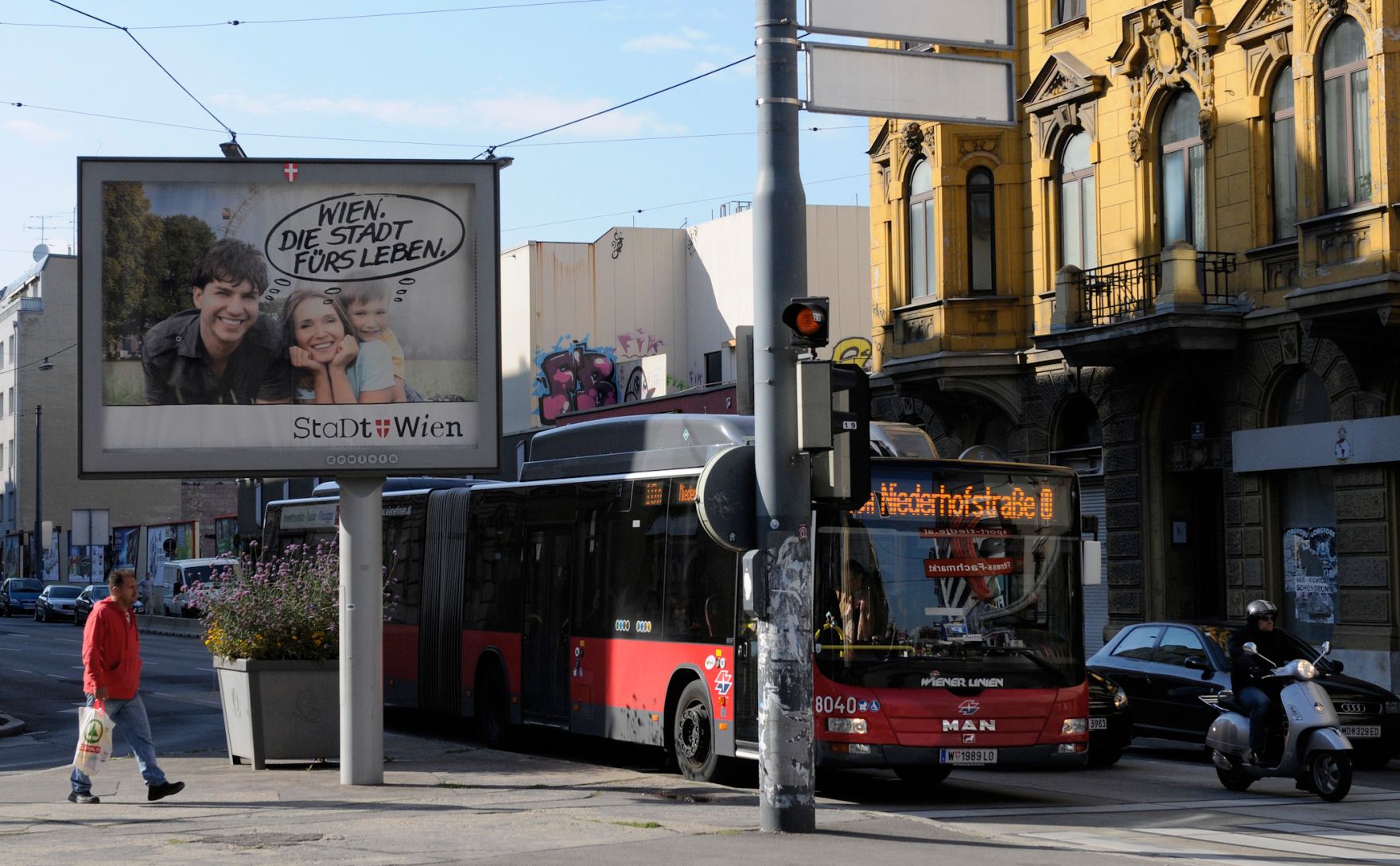 Eine Stra&szlig;enszene mit verschiedenen Verkehrsteilnehmern. Schlagworte: Autobus, Fahrzeuge, Fu&szlig;g&auml;nger, Moped, Motorrad, Person, Plakat, Stadtlandschaften, Stra&szlig;e, Verkehr, Werbung, Wiener Linien