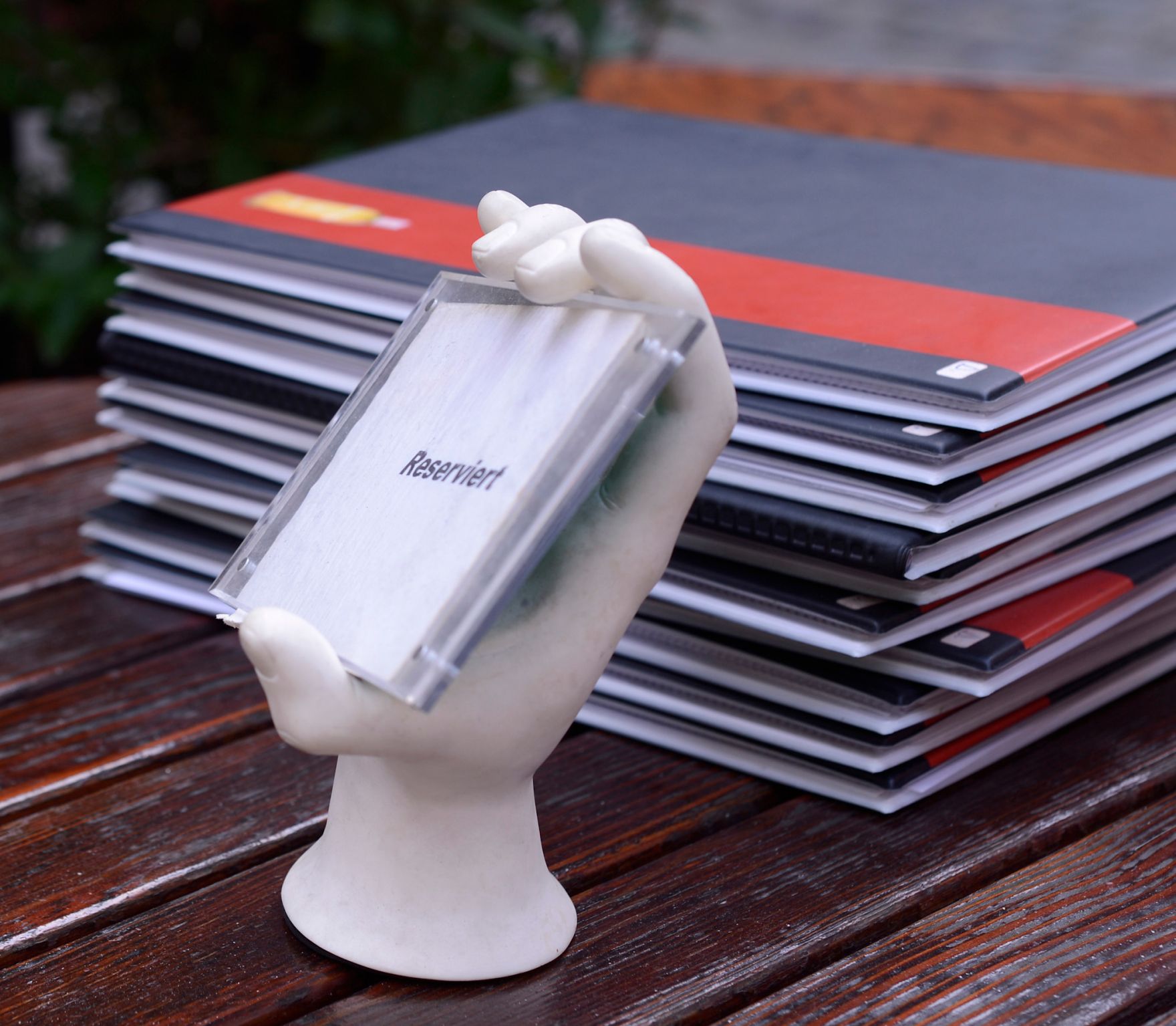 Eine Handskulptur mit einem "Reserviert"-Schild vor einem Stapel Speisekarten. Schlagworte: Beschriftung, Essen, Finger, Gastronomie, Hand, Lebensmittel, Lokal, Wirtschaft