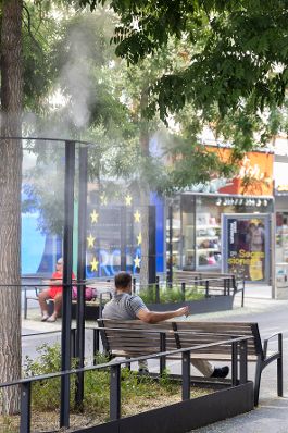 Person sitzt unter einem Baum und wird vom Spr&uuml;hnebel abgek&uuml;hlt. Schlagw&ouml;rter: Wasser, Sitzbank, Innenstadt, Baum, Spr&uuml;hnebel, EU, Europ&auml;ische Union